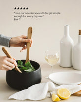 A person is serving salad from a black stoneware serving bowl on a dining table, with a white plate and a glass of white wine in the background.
